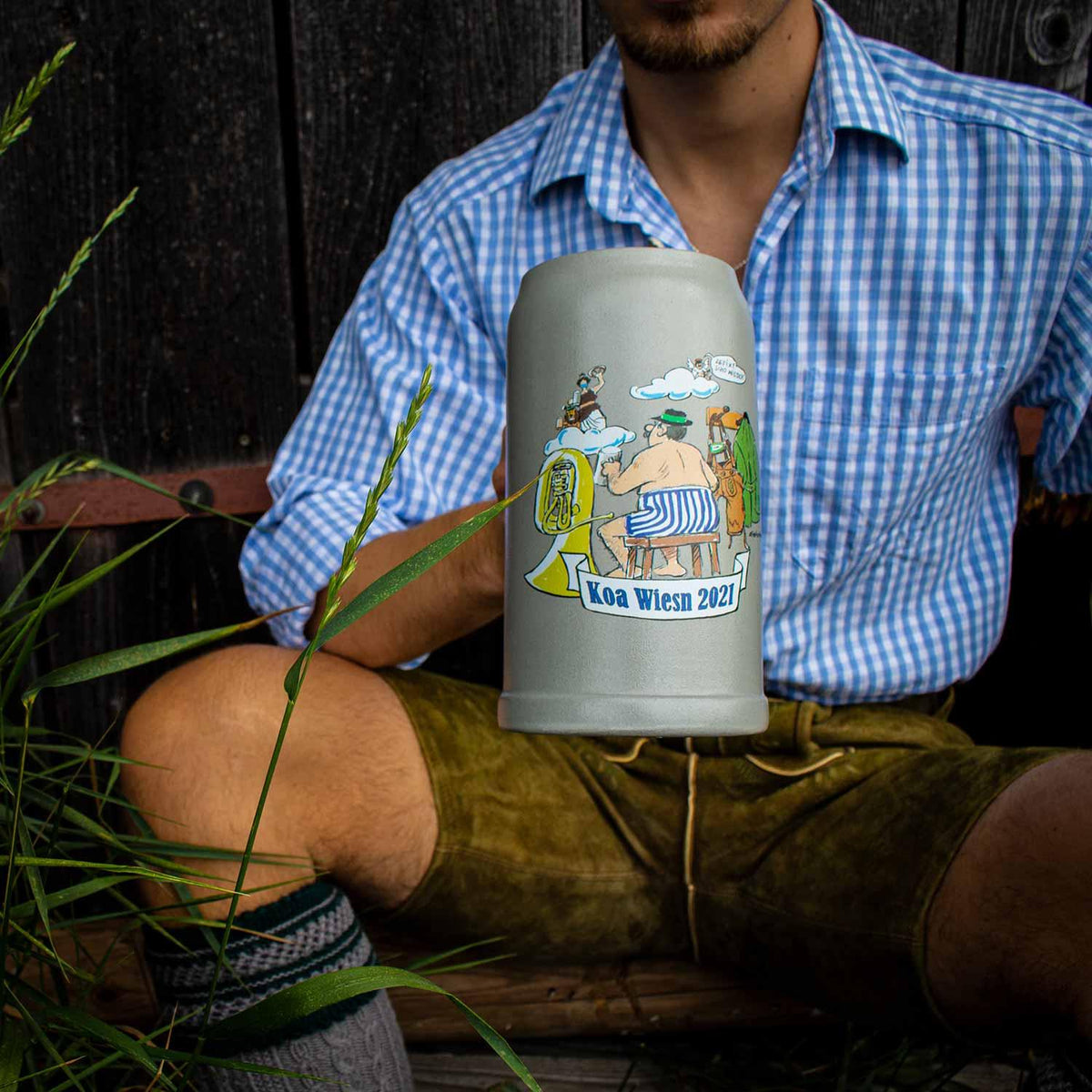 Mann in Lederhosen und kariertem Hemd hält grauen Koa Wiesnkrug 2021 mit Karikatur von Musikanten.