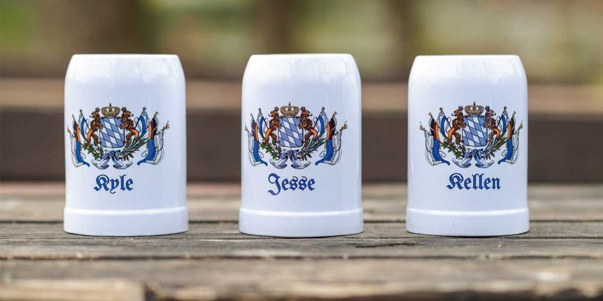 Drei weiße Keramik-Bierkrüge mit bayerischem Wappen und personalisierten Namen auf rustikalem Holztisch.
