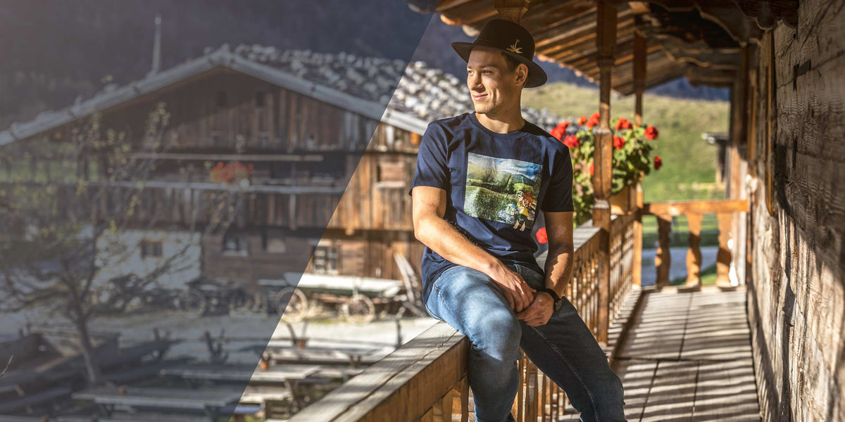 Junger Mann mit dunklem Trachtenhut und T-Shirt mit ländlichem Motiv auf rustikalem Holzbalkon.