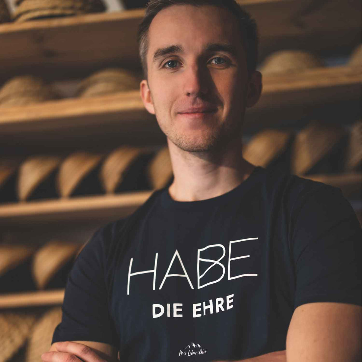 Junger Mann mit dunklem T-Shirt und dem Schriftzug "HABE DIE EHRE" vor Holzregalen mit Körben.