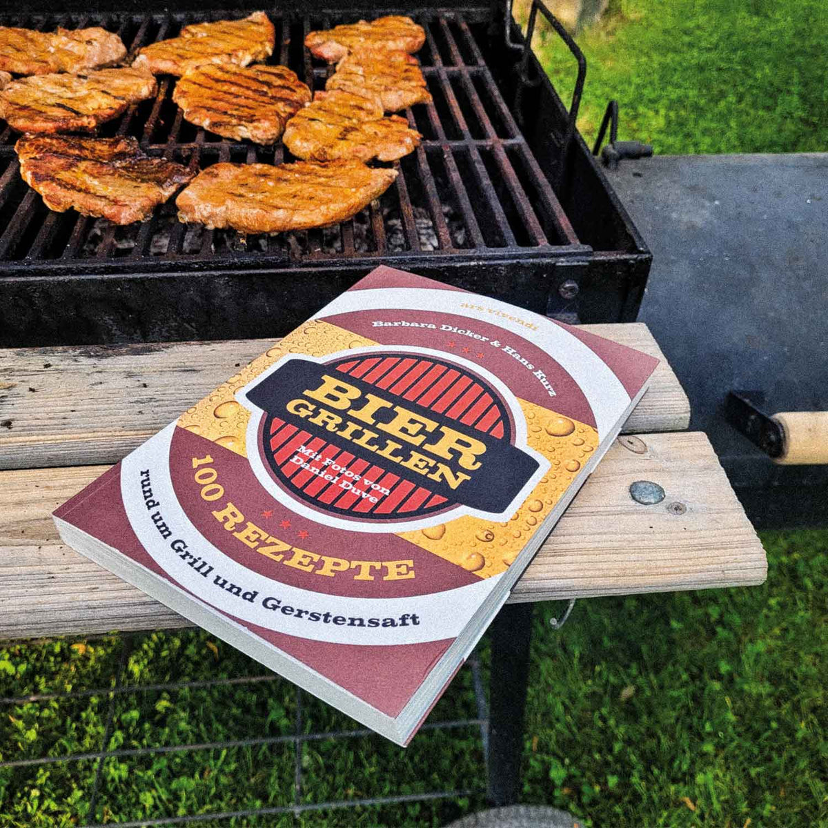 Grillbuch "Bier & Grillen" mit 100 Rezepten, neben einem Grill mit brutzelndem Fleisch im Garten.