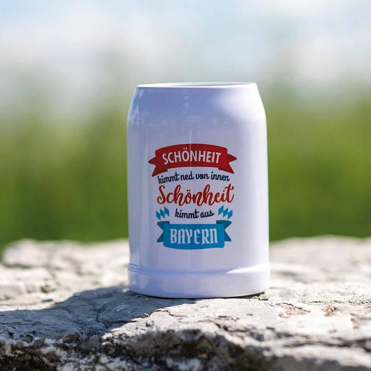 Weißer Bierkrug mit bayerischem Spruch Schöneheit kommt aus Bayern auf hellem Felsen im Freien.