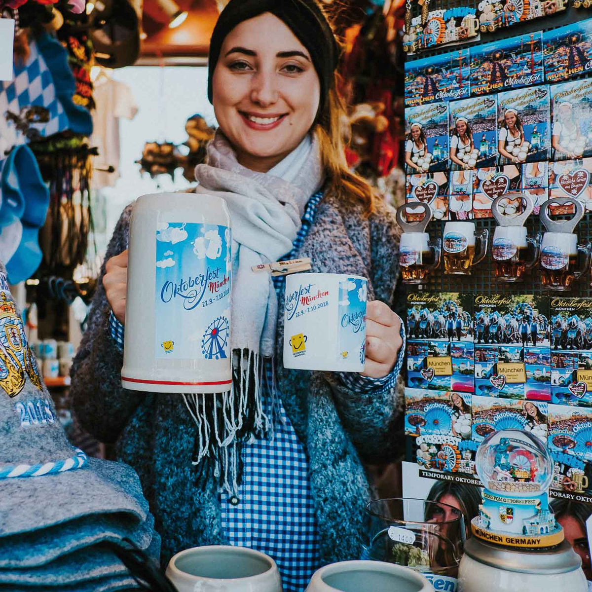 Lächelnde junge Frau präsentiert Oktoberfest-Bierkrüge beim Münchner Souvenirstand.