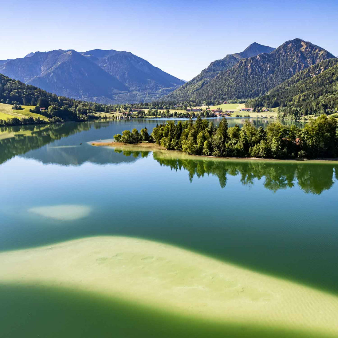 Idyllischer Bergsee mit grüner Insel, umgeben von dicht bewaldeten Bergen und Dorf in der Ferne.