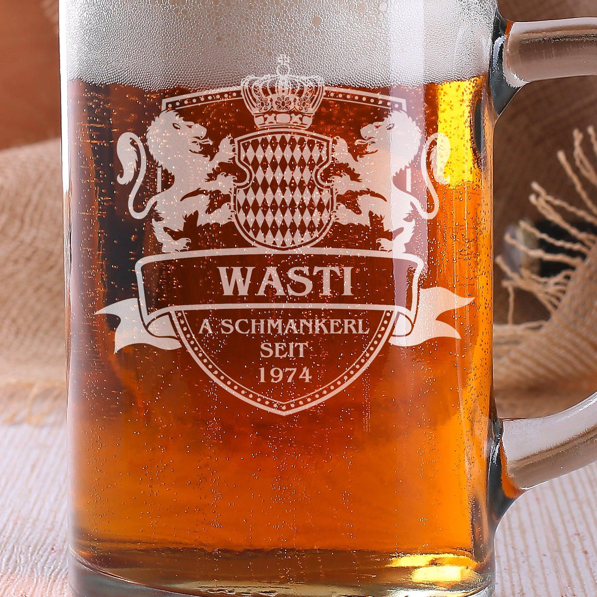 Gefüllter Bierkrug mit eingraviertem Wappen und Personalisierung "WASTI A Schmankerl SEIT 1974" auf Tisch.