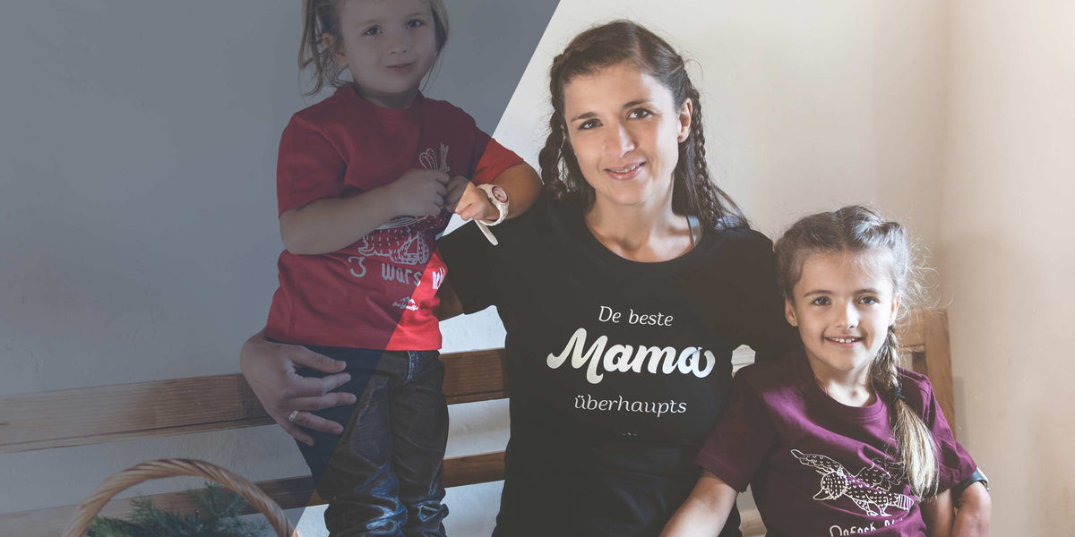 Glückliche Mutter mit geflochtenen Zöpfen im schwarzen T-Shirt "Die beste Mama überhaupt" lächelt mit ihren zwei Töchtern.