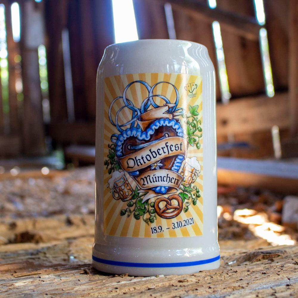 Offizieller Oktoberfestkrug 2021 mit aufgedrucktem Schriftzug München, Bierdosen und Brezel auf hellem Holz.