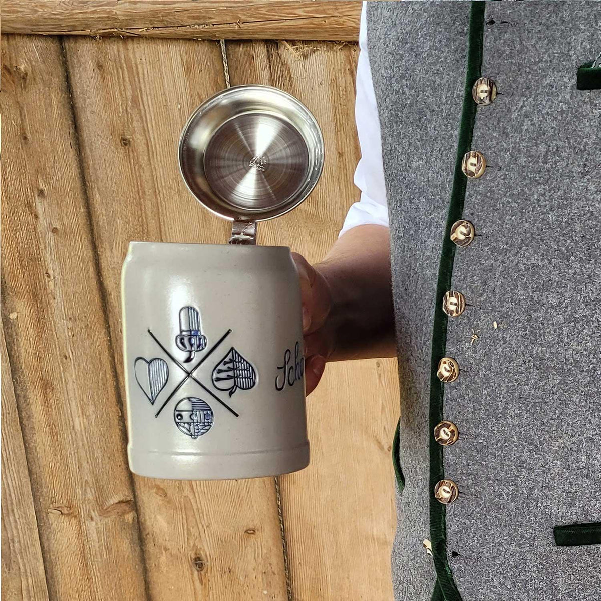 PREMIUM Bierkrug Bayrisch Blatt mit Zinndeckel, traditionellen Symbolen und "Schöner" Schriftzug in Hand eines Mannes mit Trachtenweste.