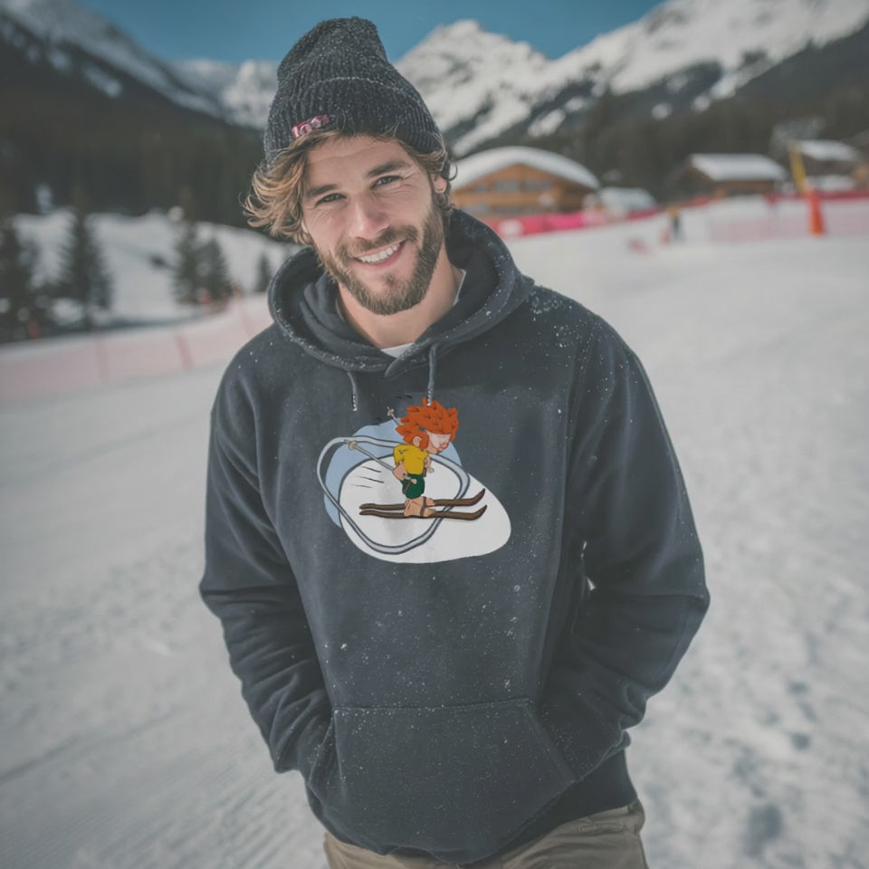 Lächelnder Mann mit Bart und Mütze im dunkelblauen Pumuckl Hoodie „Skifahren“ vor Winterlandschaft