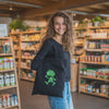 Lachende Frau mit schwarzer Stofftasche mit grünem Pumuckl Motiv in einem Naturkostladen.