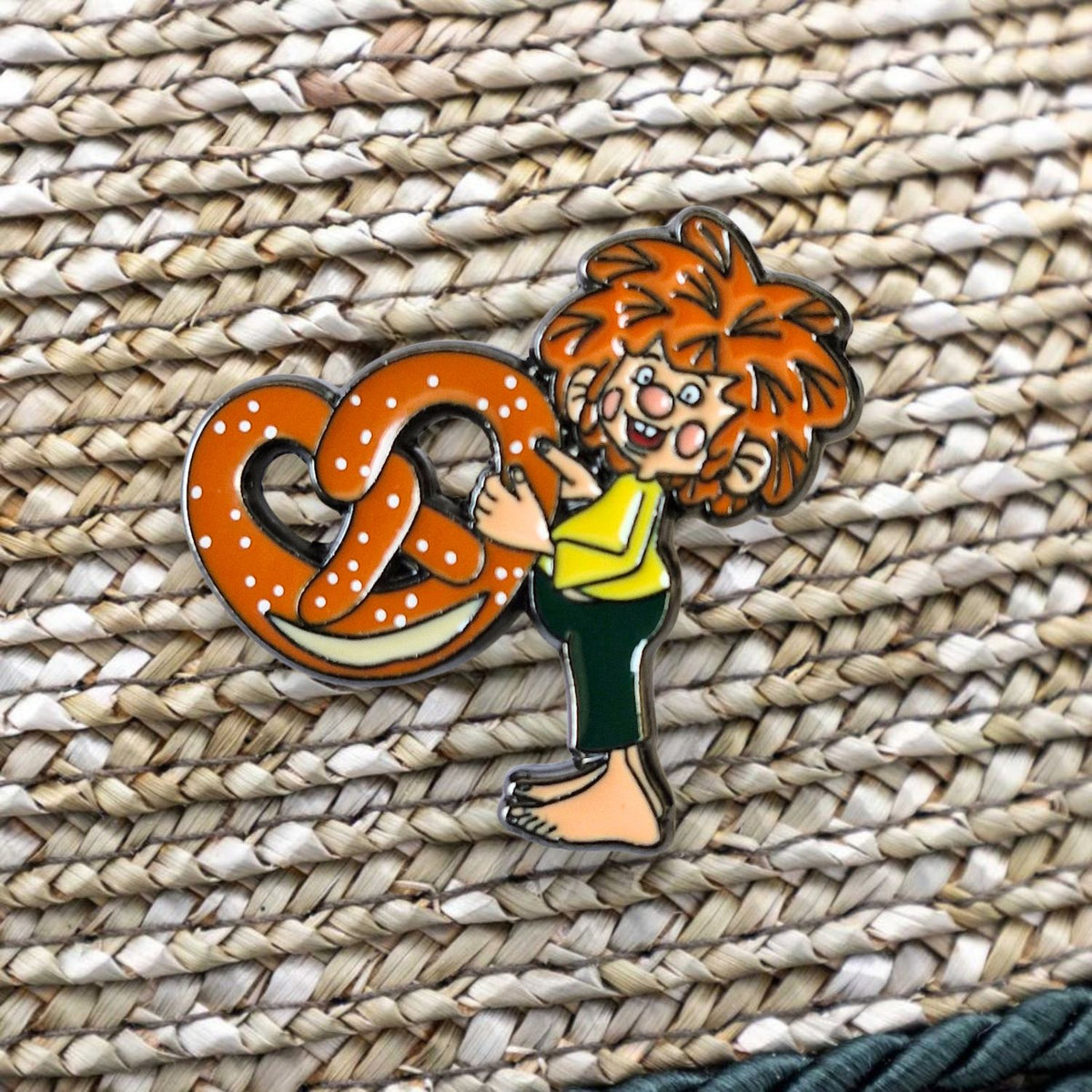 Lebhafter Pumuckl Pin mit roter Haarpracht, der eine große gesalzene Brezel umarmt, platziert auf einem geflochtenen Untergrund.
