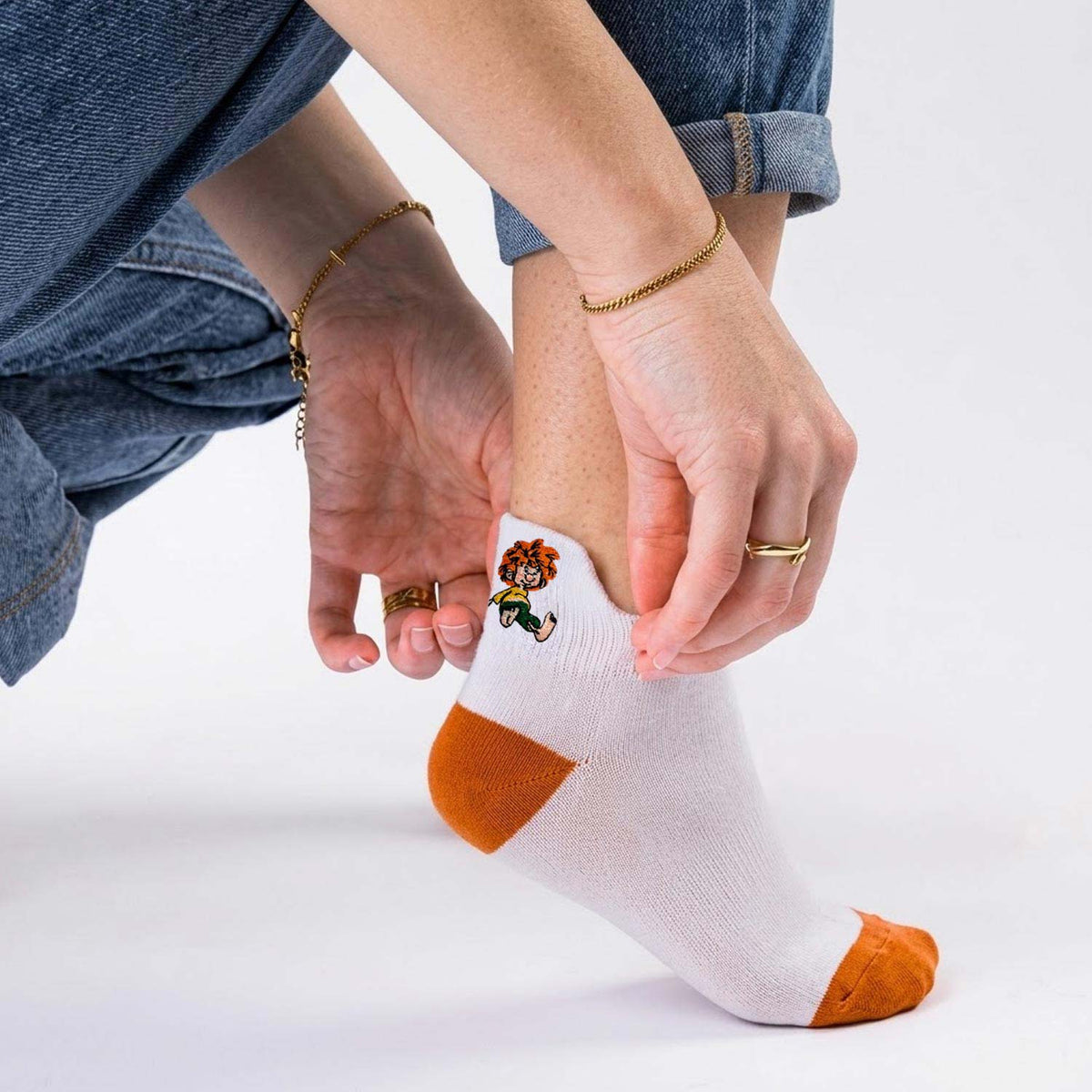 Weiße Pumuckl Sneaker Socken mit orangefarbenen Fersen- und Zehenbereichen, getragen von einer Person in Jeans.
