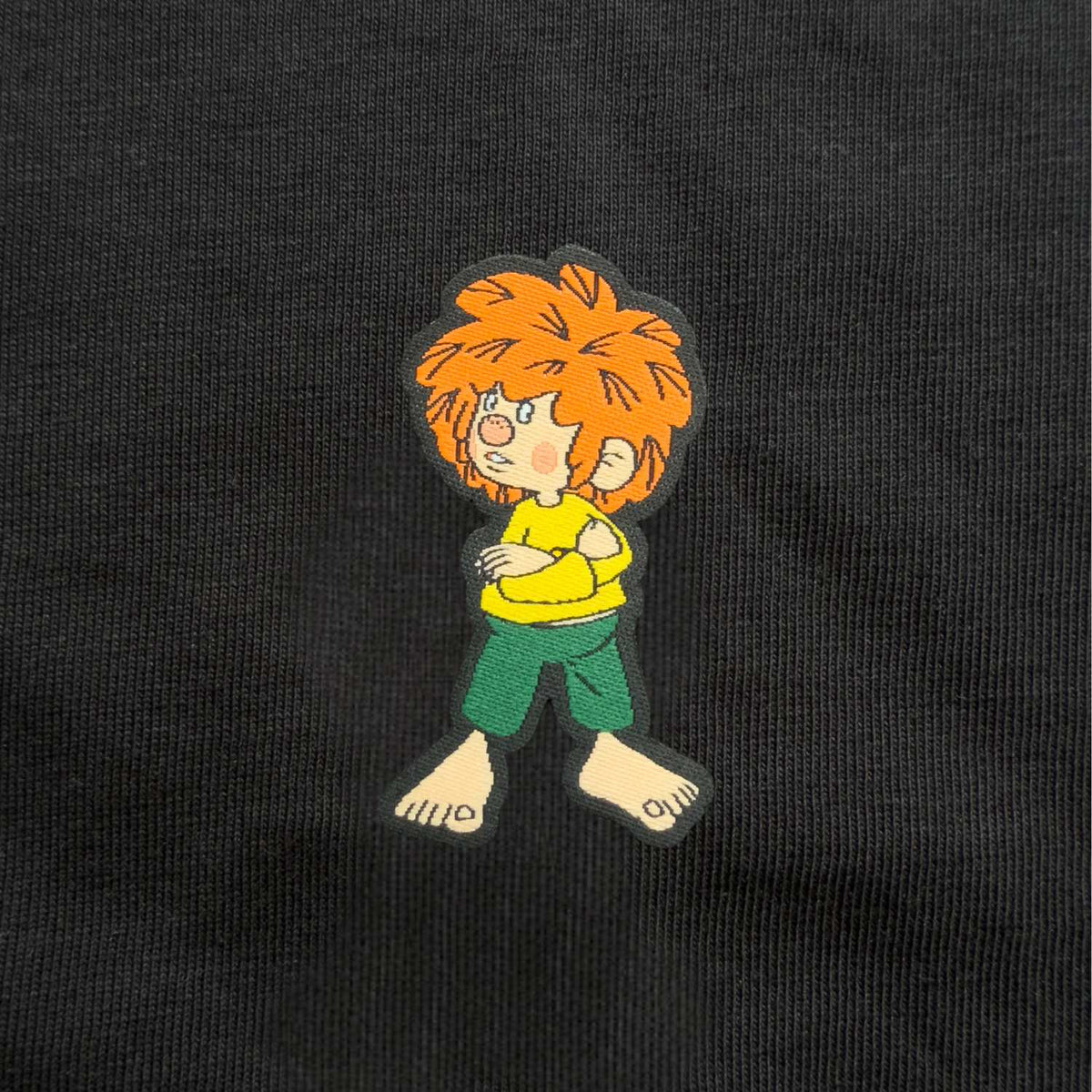 Nahaufnahme gewebtes Pumuckl-Label: grantiger Pumuckl mit verschränkten Armen auf schwarzem Damen-Shirt