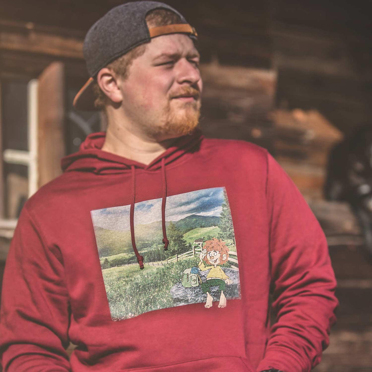 Männermodel trägt roten Pumuckl Herren Hoodie in bergiger Umgebung mit "Berg-Kobold" Motiv.