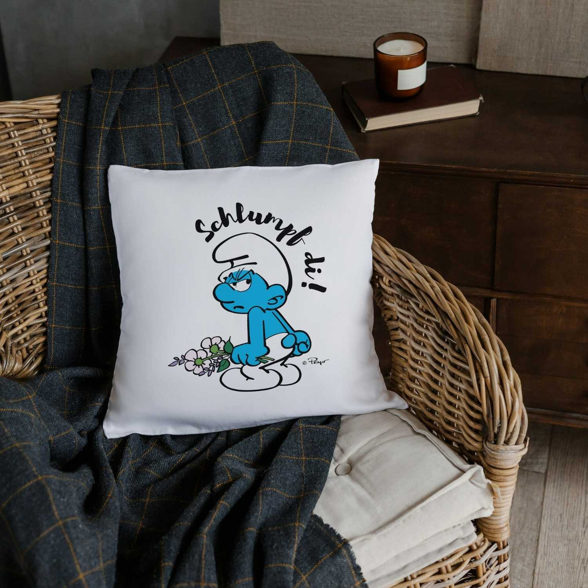 Weißes Kuschelkissen "Schlumpf di!" mit miesepetrigem Schlumpf, der Blumen hält und auf einem Rattanstuhl liegt.