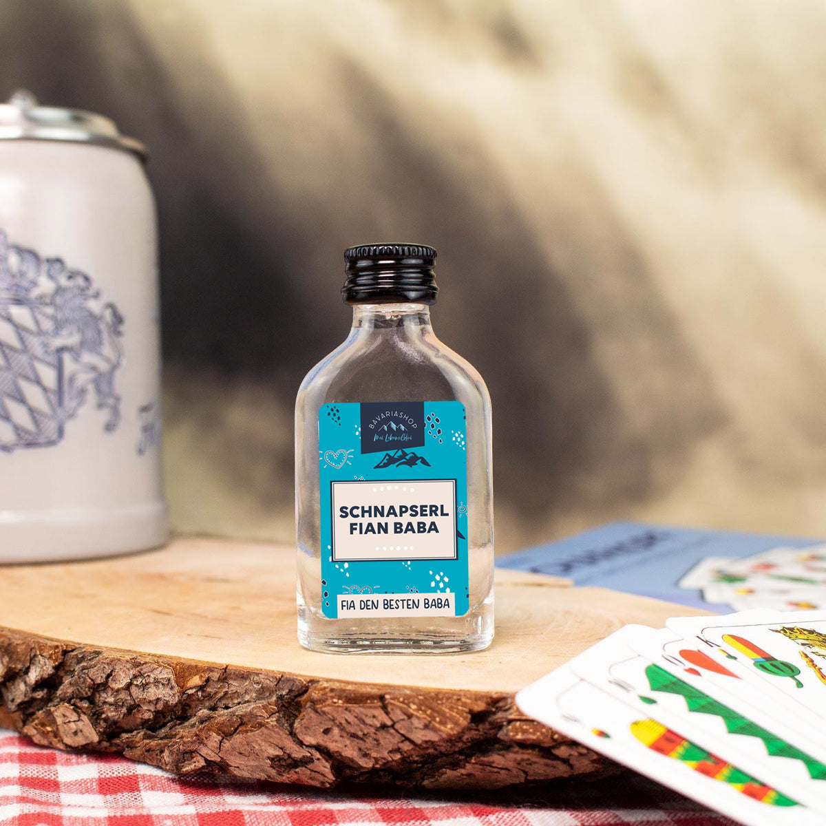Kleine Schnapsflasche "fian Baba" mit blauem Etikett auf Holzbrett, umgeben von Spielkarten und Bierkrug.