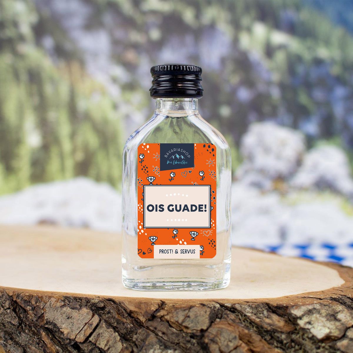 Kleines Schnapserl Ois Guade in Glasflasche mit Banderole, stehend auf rustikalem Holzbrett vor Landschaft.