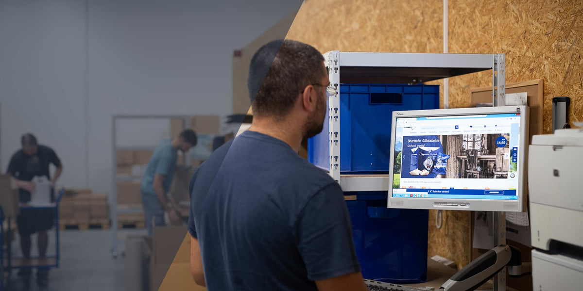 Mitarbeiter sitzt im Lager am Computer und bearbeitet Bestellungen von bayerischen Geschenkartikeln im E-Commerce-Shop.