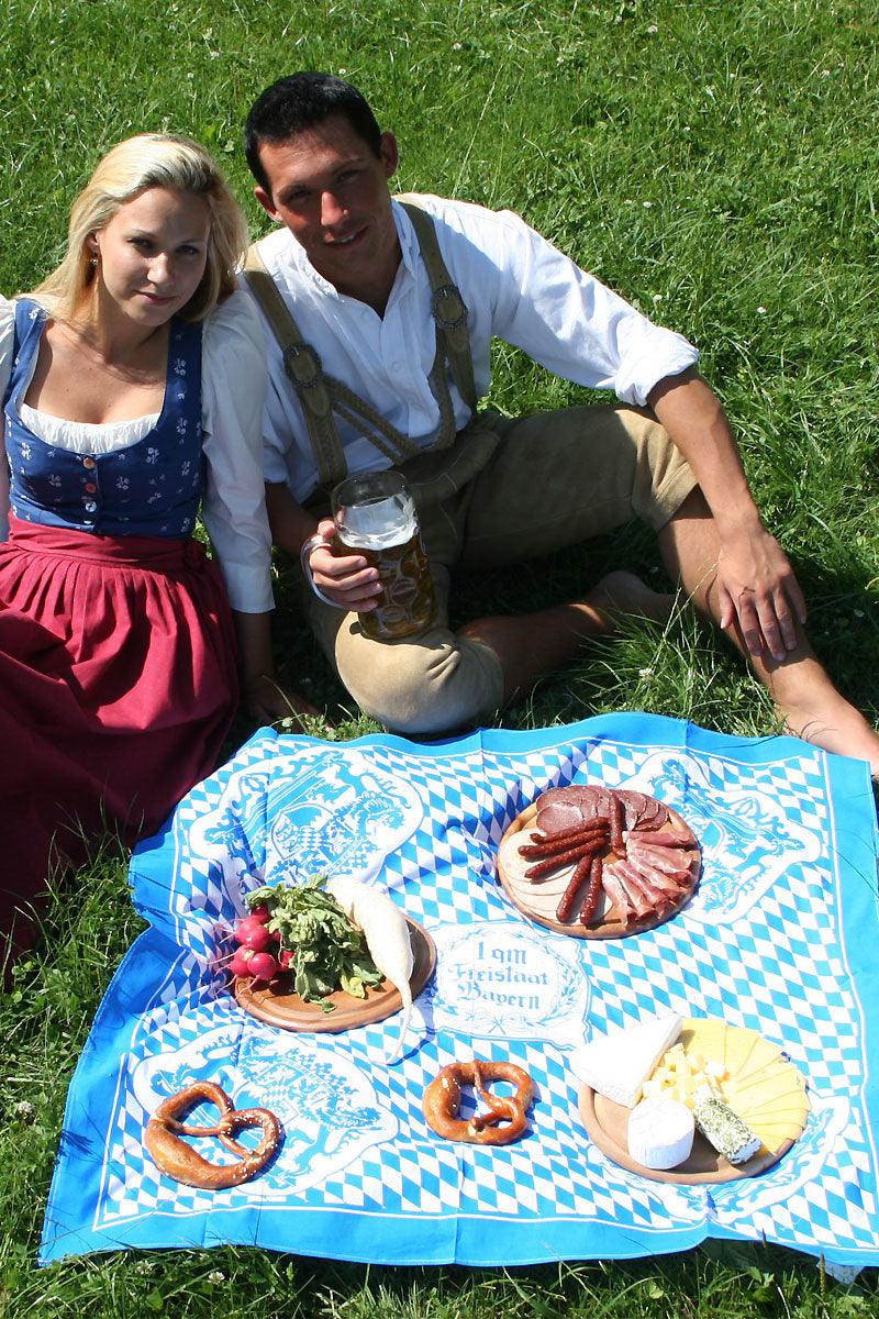 Bayerisches Paar in Tracht beim Picknick mit Brezeln, Radieschen, Wurst und Käse auf weiß-blauer Decke im Grünen.